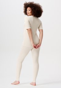 Kvinna med lockigt hår iförd en beige långärmad tröja och leggings, står barfota och ser över axeln med händerna knäppta bakom ryggen.