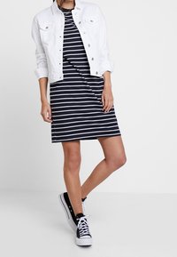 Randig marinblå klänning med vita horisontella ränder, matchad med en vit denimjacka. Bärs med svart och vit sneakers.