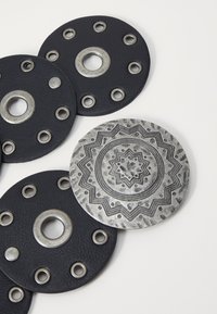 Cirkulāru aksesuāru komplekts, kas ietver divus melnus ādas gabalus ar metalizētiem caurumiem un sudraba teksturētu disku ar sarežģītu rakstu.