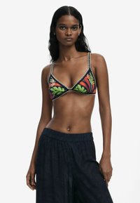 Schwarzes Floral-Bikini-Oberteil mit lebhaften grünen und orangen Blättern, verziert mit dekorativen Nähten entlang der Ränder. Das Model trägt schwarze gemusterte Hosen.