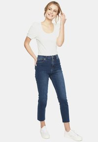 Biała bluzka z krótkim rękawem i okrągłym dekoltem, połączona z ciemnoniebieskimi spodniami na wysokim stanie. Modelka ma na sobie białe trampki i stoi na neutralnym tle.