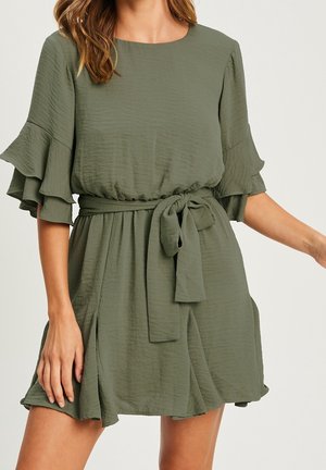 Kvinde iført en olivengrøn kjole med korte ærmer, flæser på ærmerne og et bæltet bånd bundet om livet.