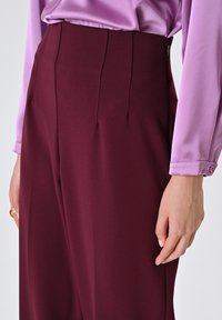 Pantaloni bordeaux a vita alta con pieghe frontali nette, abbinati a una blusa di raso lilla con polsini bottonati.