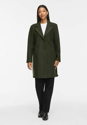 Mujer con un abrigo verde oscuro abotonado sobre una camiseta blanca, pantalones negros y botines negros, de pie frente a un fondo blanco liso.