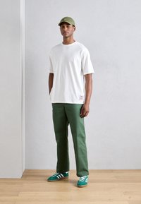 Camiseta de algodón blanca, pantalones de algodón verdes y gorra verde. Los zapatos son de gamuza verde con rayas blancas y suela de goma. Fondo simple.