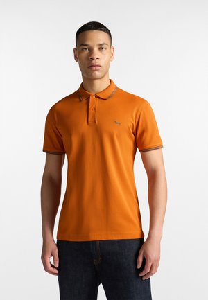 Camisa polo naranja con una tapa de tres botones, costuras de acento azules, mangas cortas y un pequeño logo en el lado izquierdo del pecho. Combinada con jeans oscuros.