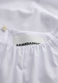 Vêtement blanc avec un col froncé et une étiquette en tissu cousue visible montrant partiellement le texte « ARMEDANGEL ».