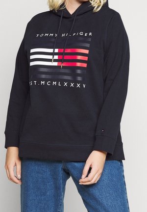 Person trägt schwarzen Tommy Hilfiger Hoodie mit Logo und Streifen in Weiß, Rot und Schwarz, kombiniert mit blauen Jeans, steht mit entspannten Händen.