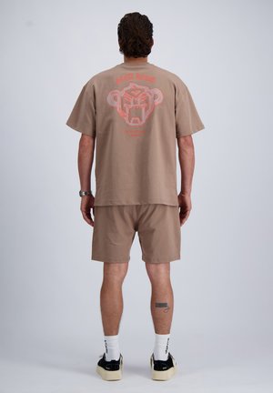Man draagt beige short en t-shirt met rood abstract gezicht op de rug, witte sokken, zwart-witte sneakers, staat met de rug naar de camera gekeerd.