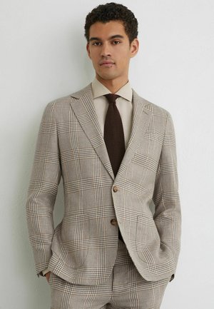 Muž v béžovom kockovanom obleku, s krémovou košeľou a tmavohnedou kravatou, stojaci s rukami v kapse pri bielej stene.