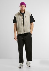 Beige fleece vest with a high collar, two zip pockets, worn over a black t-shirt, paired with black pants and layered sneakers.