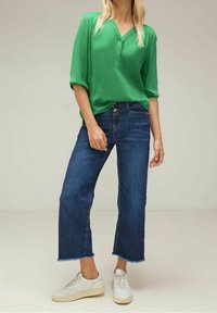 Chemisier vert à boutons et manches courtes, associé à un jean en denim bleu à jambes larges avec des ourlets effilochés. Des baskets blanches complètent la tenue.