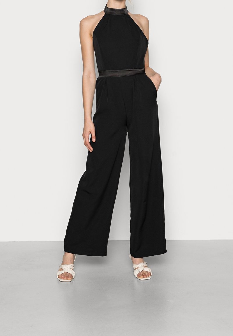 Combinaison noire sans manches avec jambes larges, présentant un corsage ajusté et une ceinture en satin. Le modèle porte des sandales à strap torsadé beige clair.