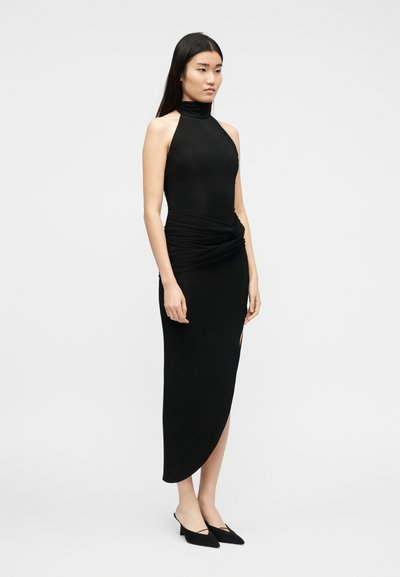 Γυναίκα που φοράει μαύρο αμάνικο φόρεμα με ψηλό λαιμό, με ζώνη στη μέση και ασύμμετρο τελείωμα, φορώντας μαύρα μυτερά τακούνια.