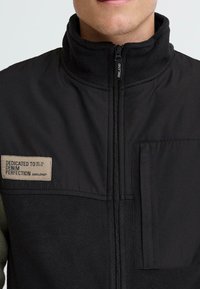 Schwarze Jacke mit Fleece-Textur, Reißverschluss vorne, hohem Kragen und einer Brusttasche. Mit einem Etikett, das "Engagiert für Denim-Vollkommenheit" besagt.