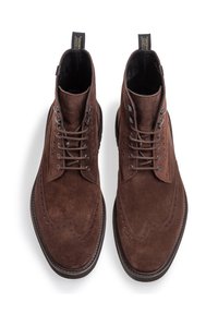 Stivaletti tronchetti in suede marrone con chiusura lacci, dettagli wingtip e suola texturizzata, dotati di un'asola sul retro per un facile indosso.