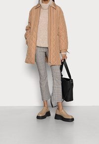 Giacca beige trapuntata con colletto alto, maglione lavorato a maglia crema, pantaloni scozzesi e scarpe slip-on beige con zeppa. Borsa tote nera.