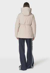 Chaqueta acolchada beige con cintura ajustada, cuello alto y capucha. Combinada con pantalones de mezclilla oscuros de pierna ancha que presentan franjas grises en los costados.