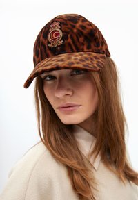 Cappello a motivo leopardo realizzato in tessuto morbido. Presenta un logo ricamato sulla parte frontale. Schema di colori marrone e beige con visiera curva.