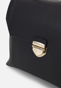 Borsa a mano in pelle nera con patta curva, caratterizzata da una chiusura in metallo dorato. Il design è elegante con accenti minimalisti e una finitura strutturata.