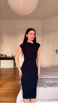 Γυναίκα με μακριά σκουρόχρωμα μαλλιά, φορώντας εφαρμοστό μαύρο αμάνικο φόρεμα, στέκεται σε ένα υπνοδωμάτιο με ξύλινο δάπεδο και λευκούς τοίχους.