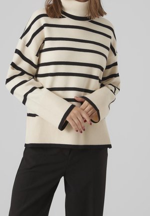 Femme portant un pull à col roulé crème avec des rayures noires horizontales et un pantalon noir, mains jointes à la taille.
