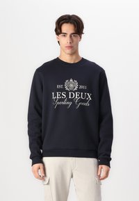 Sudadera azul marino con cuello redondo, mangas largas y puños acanalados. Presenta texto de logo impreso en blanco y el año de establecimiento en la parte frontal.