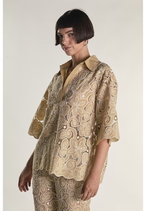 Kvinde med kort mørkt hår bærer beige blomstret blondebluse og matchende bukser mod ensfarvet baggrund, kigger let til siden.