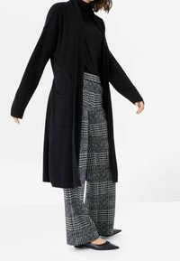Lång svart kofta med öppen front, stickad textur och sidofickor, tillsammans med grå- och svartrutiga vida byxor och svarta spetsiga skor.