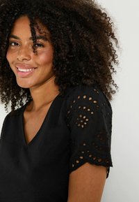 Top negro de cuello en V con intrincadas mangas cortas de encaje de ojos, textura suave y un ajuste relajado, que acentúa un diseño moderno.