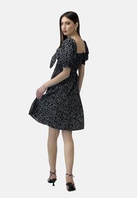 Vestido negro con flores, con mangas cortas abullonadas, escote cuadrado y silueta ajustada en la parte superior y acampanada en la parte inferior, combinado con sandalias de tiras negras.