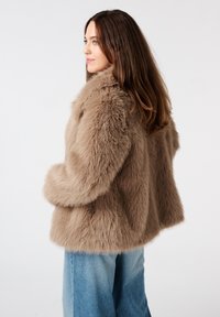 Femme aux longs cheveux bruns portant un manteau en fourrure beige moelleux et un jean bleu, tournée vers la gauche sur un fond blanc uni.