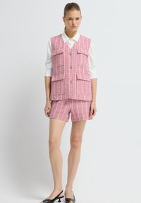 Růžová a bílá tkaná vesta se dvěma kapsami a zlatými knoflíky, spárovaná s odpovídajícími šortkami. Modelka má na sobě bílou košili a černé podpatky.