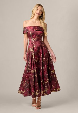 Blondine in een bordeauxrode bloemenjurk met blote schouders en gouden accenten, staand tegen een effen lichte achtergrond.