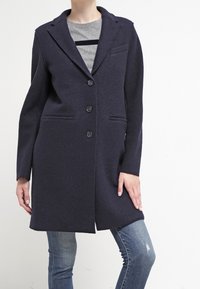 Tummansininen villakangastakki, jossa on läpälliset kaulukset, yksirivinen malli, kolme nappia ja sivutaskut, puettuna harmaan paidan ja farkkujen päälle.