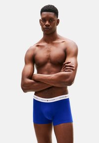 Calzoncillos bóxer de algodón azul con una cintura blanca con la marca, diseño ajustado y textura suave, usados por un modelo masculino con los brazos cruzados.