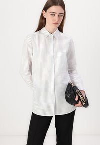 White button-up shirt with vertical black stripes, long sleeves, and a chest pocket. Model holds a small black quilted handbag.