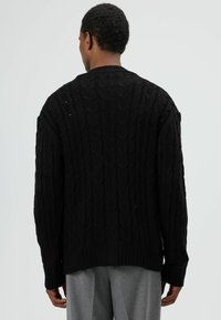 Zwarte gebreide cardigan met een kabelpatroon, geribbelde manchetten en zoom, losse pasvorm en een hoge kraag, gedragen over lichtgrijze broek.