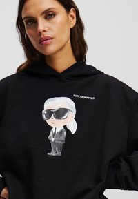 Sweat à capuche noir avec une illustration cartoon d'une silhouette portant des lunettes de soleil et un costume. Du texte "KARL LAGERFELD" imprimé au-dessus du design.