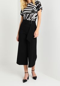 Czarne spodnie culotte z paskiem w talii, zestawione z czarno-białą kreszową bluzką z wzorem liści. Czarne buty na obcasie.