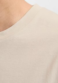 Beżowa bawełniana koszulka z okrągłym dekoltem, charakteryzująca się gładką teksturą i delikatnym przeszyciem wokół dekoltu. Zbliżenie na tkaninę.