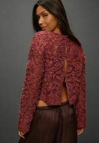 Lace crop top in deep burgundy with floral appliqué, long sleeves, open back, and a tied closure. Paired with smooth brown pants.
