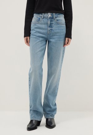 Persona con jeans azul claro de pierna recta, botas negras con tacón y punta cuadrada, y una blusa negra de manga larga, de pie contra un fondo liso.