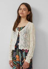 Kantgebreide cardigan in off-white met een cropped ontwerp, voorzien van een striksluiting en geschulpte randen, gedragen over een bloemenpatroonjurk.
