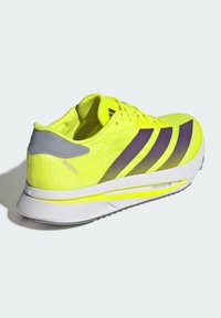 Chaussure de course néon jaune avec des accents violets et gris, partie supérieure en maille, col de talon rembourré et semelle intermédiaire blanche, conçue pour la performance et le confort.