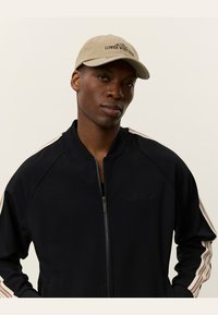 Black zip-up jacket with striped brown accents on sleeves. Worn with a beige cap featuring "Lower West Side" text.