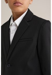 Czarna, dopasowana marynarka wykonana z fakturowanej tkaniny, z jednym guzikiem, klapami z wycięciem i kieszenią na piersi. Pod spodem biała koszula z kołnierzykiem.