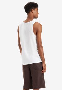 Top bianco senza maniche con scollatura arrotondata, realizzato in cotone. Abbinato a pantaloni corti marroni fino al ginocchio con tasche sul retro e dettagli cuciti.
