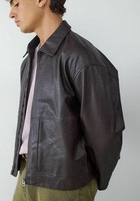 Bruine leren jas met een kraag, ritssluiting en gestructureerde schouders. Glad oppervlak met zichtbare stikseldetails.