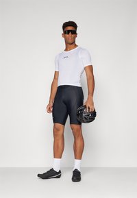 Un maillot blanc à manches courtes avec une texture en mesh, associé à un short noir ajusté. Des chaussures de cyclisme noires et un casque noir à la main.